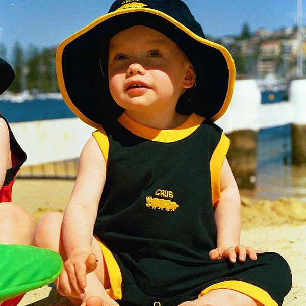 a baby sitting on the beach wearing ozi varmints toweling hat